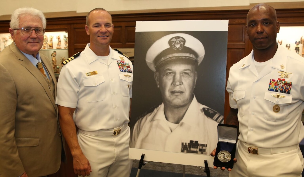 Two men in military dress uniforms and a man in a suit pose for a photo with a blown-up photo of a sailor wearing a dress uniform.