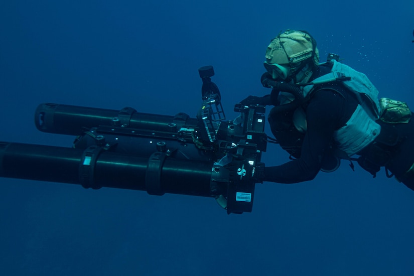 A scuba diver pushes a large black apparatus through deep blue water.