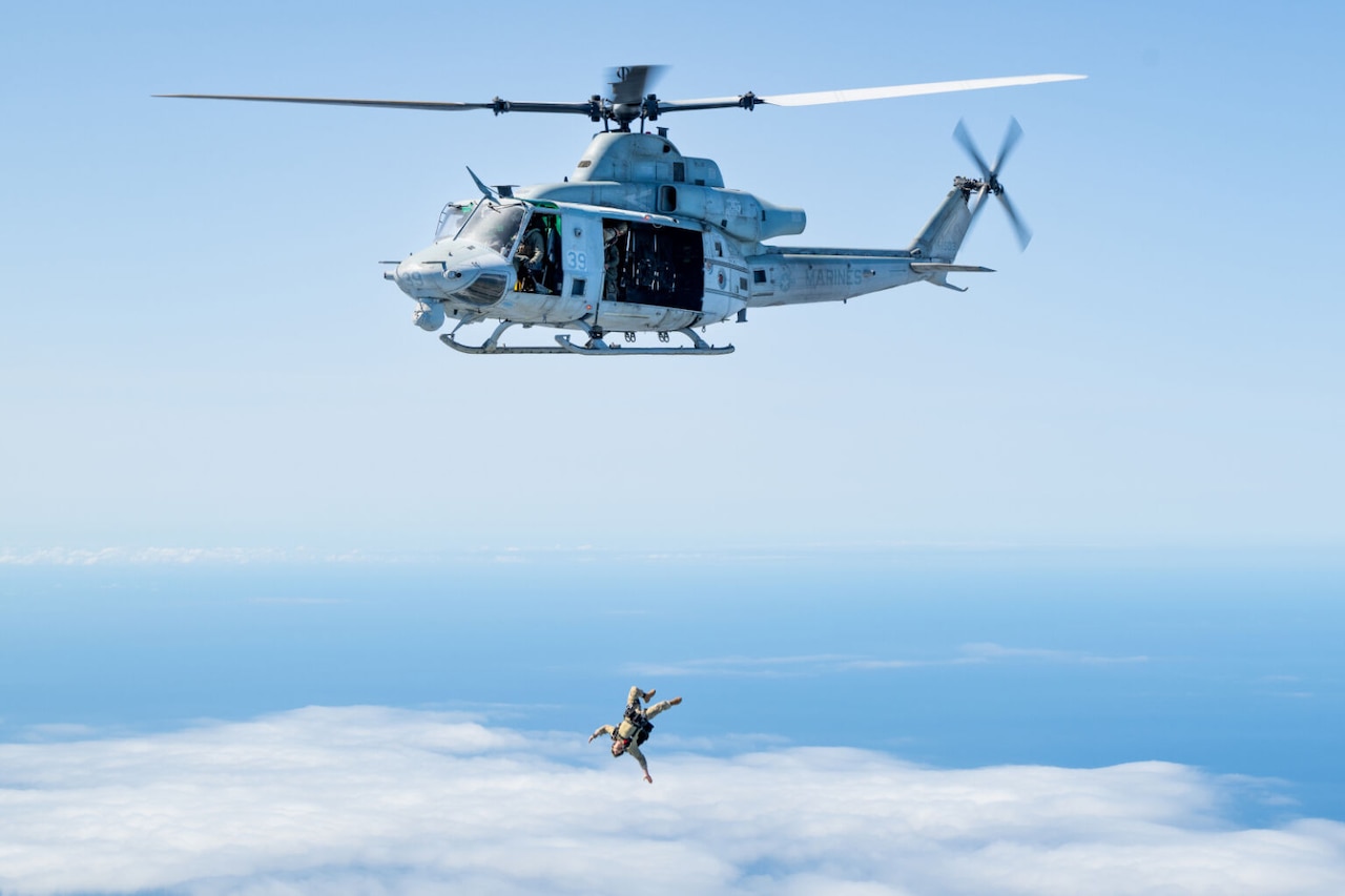 A skydiver in a military uniform free falls below a helicopter flying in the blue sky.
