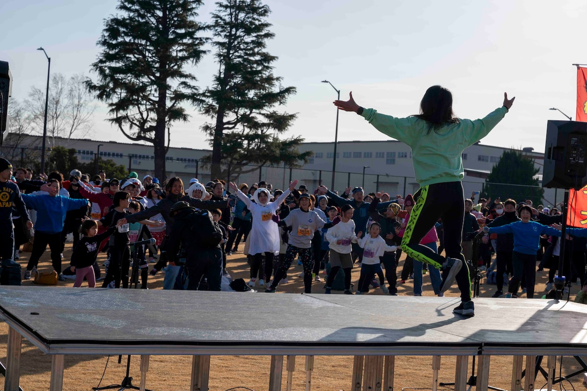 A woman does warm up exercises on a stage as a crowd follows along.