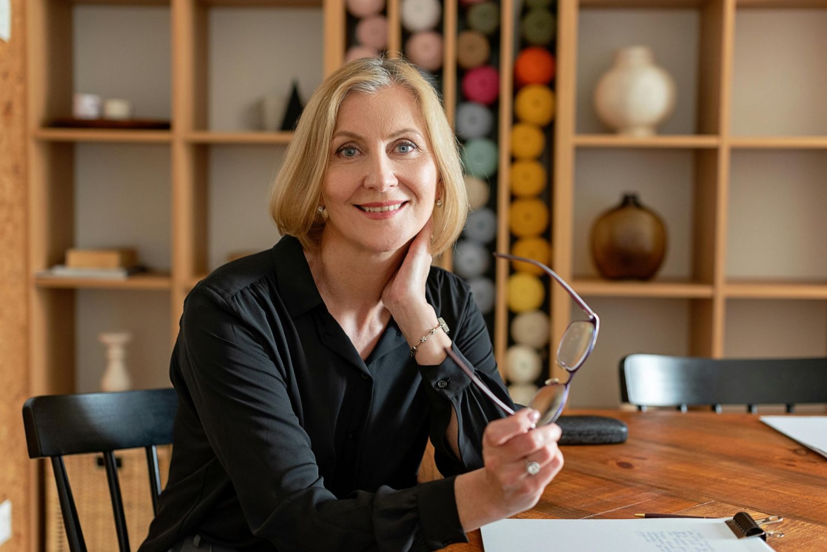 Smiling woman sits at a table while holding her glasses.