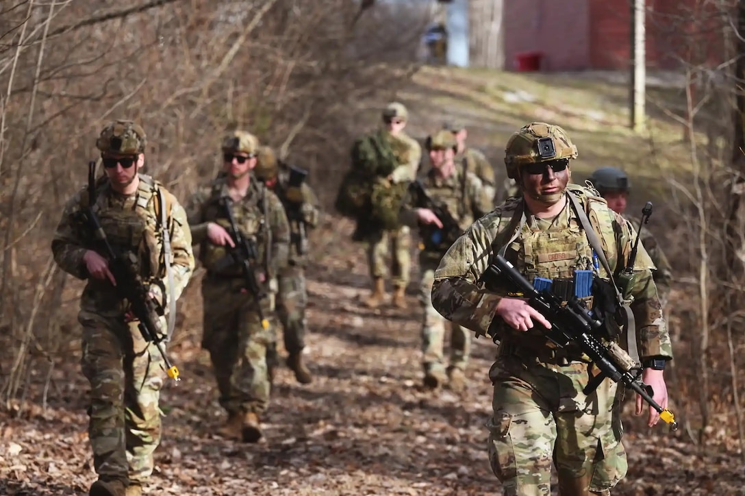 Seven members of the US military in uniforms and carrying weapons patrol a footpath next to some trees.