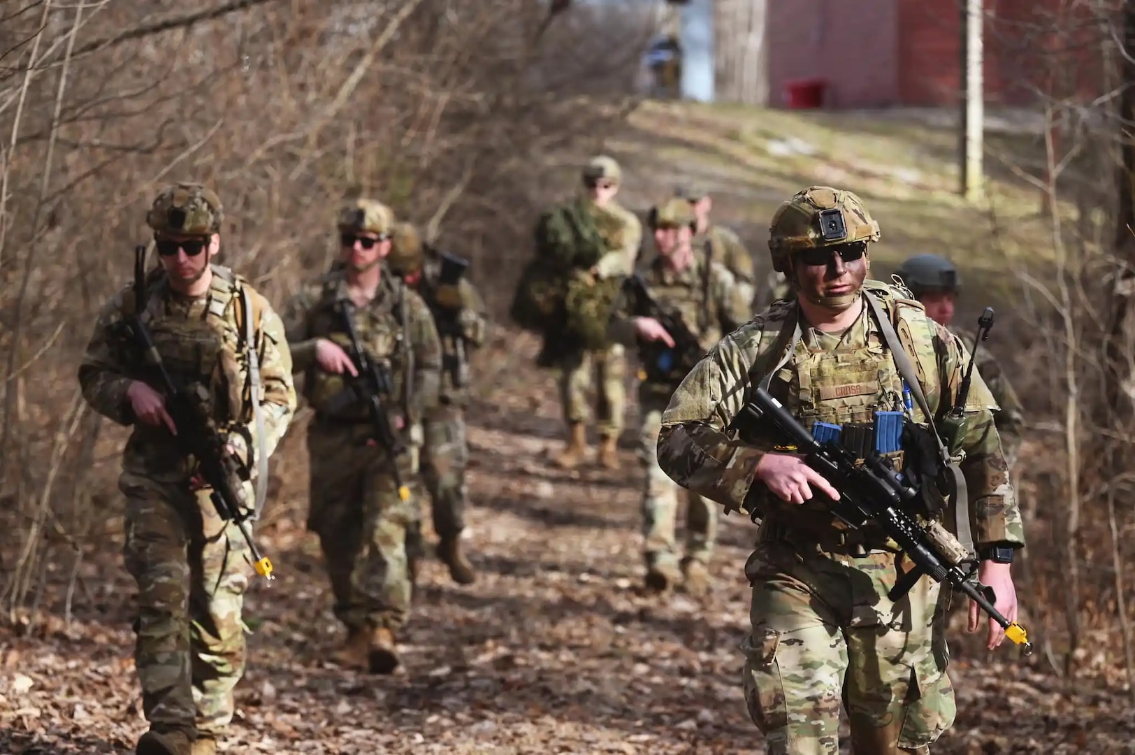 Seven members of the US military in uniforms and carrying weapons patrol a footpath next to some trees.