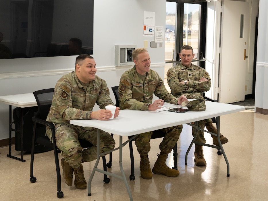 Three Airmen in camouflage uniforms sitting at a white folding table inside a room with large windows and a door.