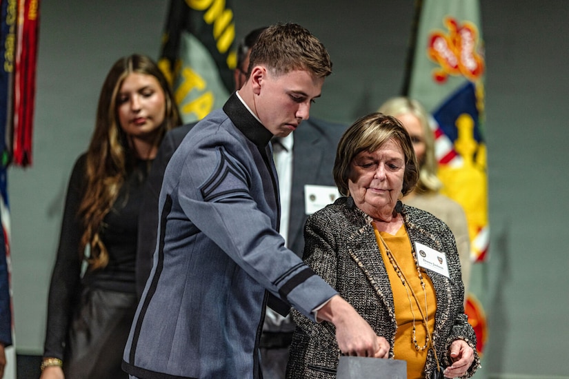 A man in a military dress uniform and an older woman in business attire place gold rings into a crucible sitting on a table. There are two women and a man in business attire standing behind them.