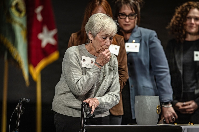 An older woman in casual attire kisses a ring in her hand as she stands on stage. The woman is looking at a crucible on a table in front of her. There are three women in business attire standing in line behind her.