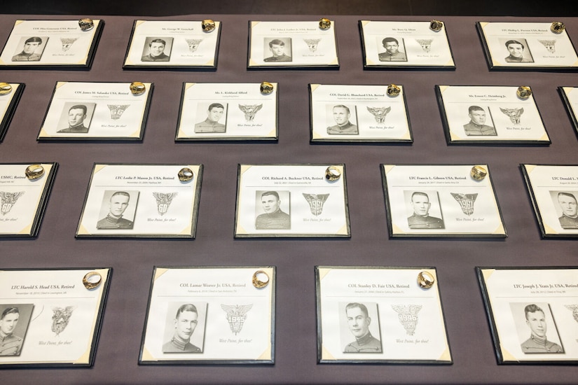Black and white photos of men in military dress uniforms are displayed in rows on a table as gold rings sit on top of each photo.