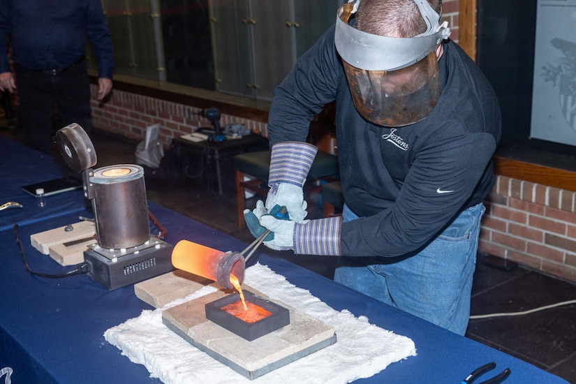 A man in casual attire with gloves and a protective visor on his face pours melted gold into a mold on a table.