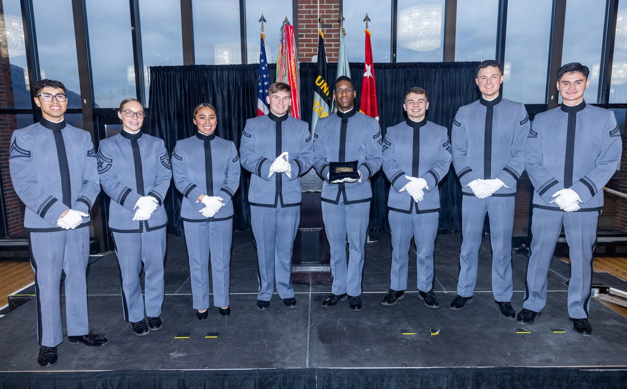 Six men and two women in military dress uniforms stand in a line on a stage as they pose for a photo. Several flags hang in the background.