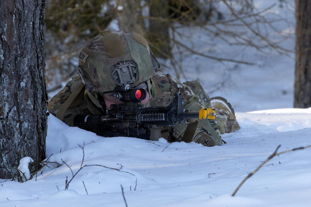 A person lying on their stomach outdoors in the snow wearing a camouflage military uniform, helmet and goggles looks through the scope of a weapon; the outer end of the scope is red.