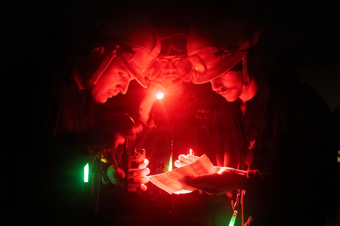 Three men wearing military uniforms and helmets with goggles on top read a checklist at night; one holds a pen and paper; another shines a small flashlight downward; another holds a communication device.