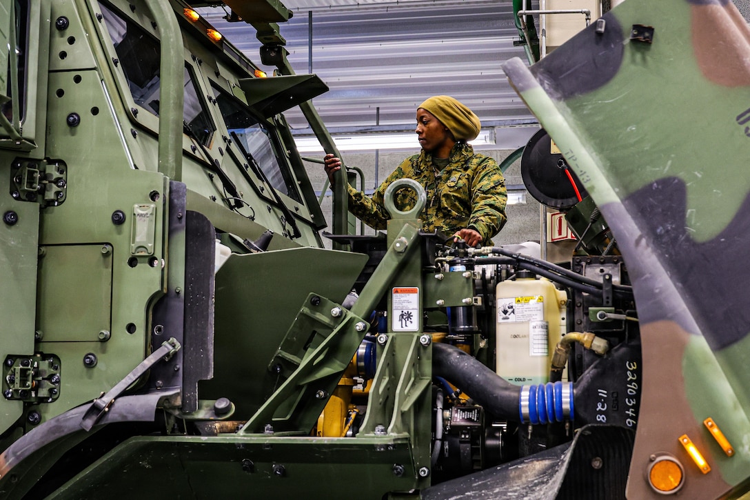 A woman wearing a camouflage military uniform and head cover stands above a large truck motor for inspection.