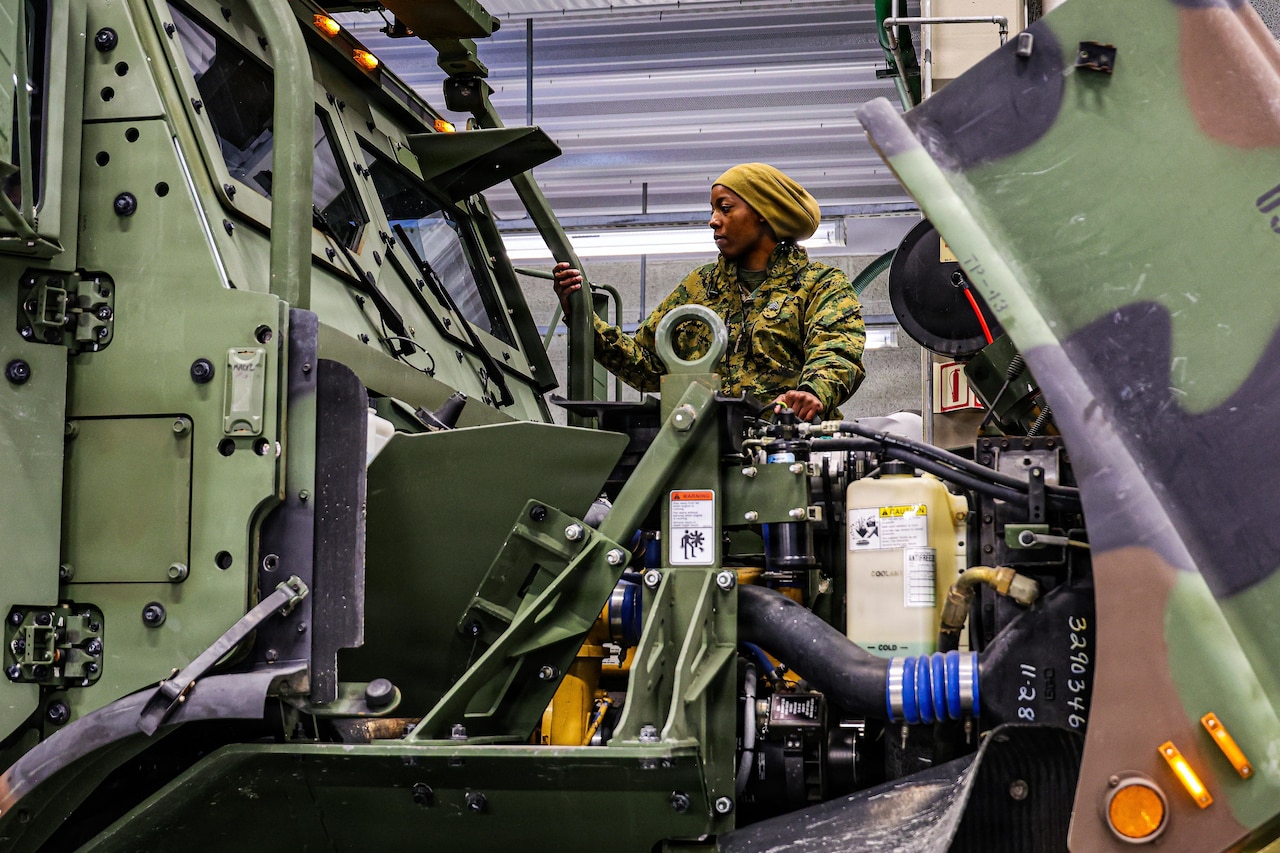 A woman wearing a camouflage military uniform and head cover stands above a large truck motor for inspection.