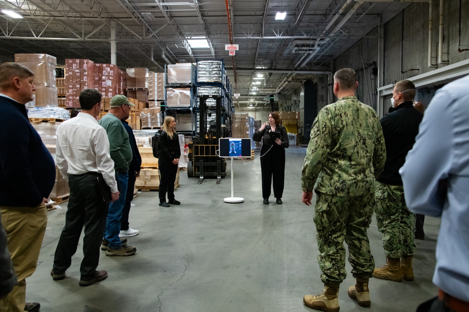 A group of people stand inside a warehouse talking to each other and looking at two people wearing lidar backpacks and pointing to a display screen.