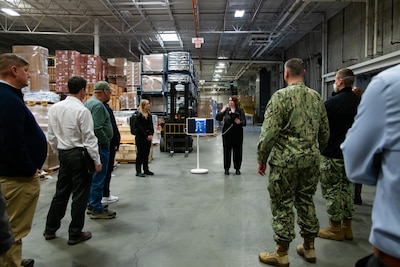 A group of people stand inside a warehouse talking to each other and looking at two people wearing lidar backpacks and pointing to a display screen.