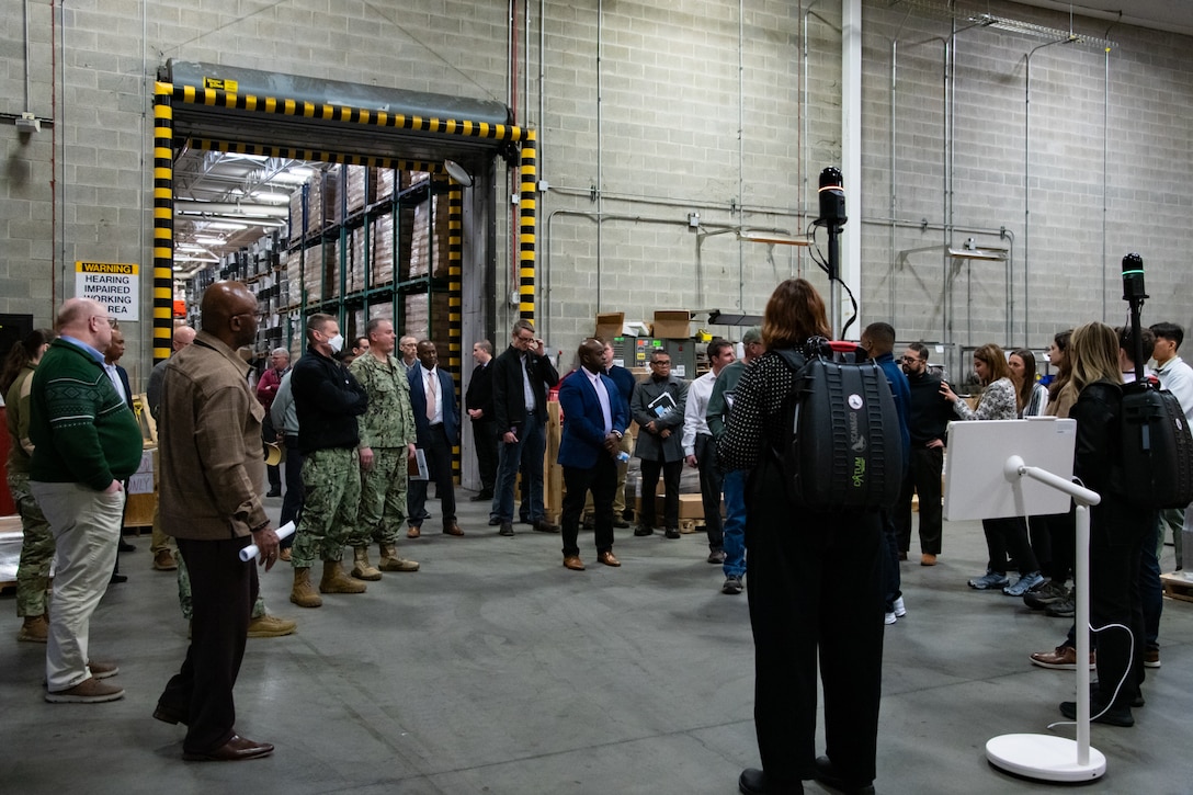 A group of people stand inside a warehouse talking to each other and looking at two people wearing lidar backpacks.