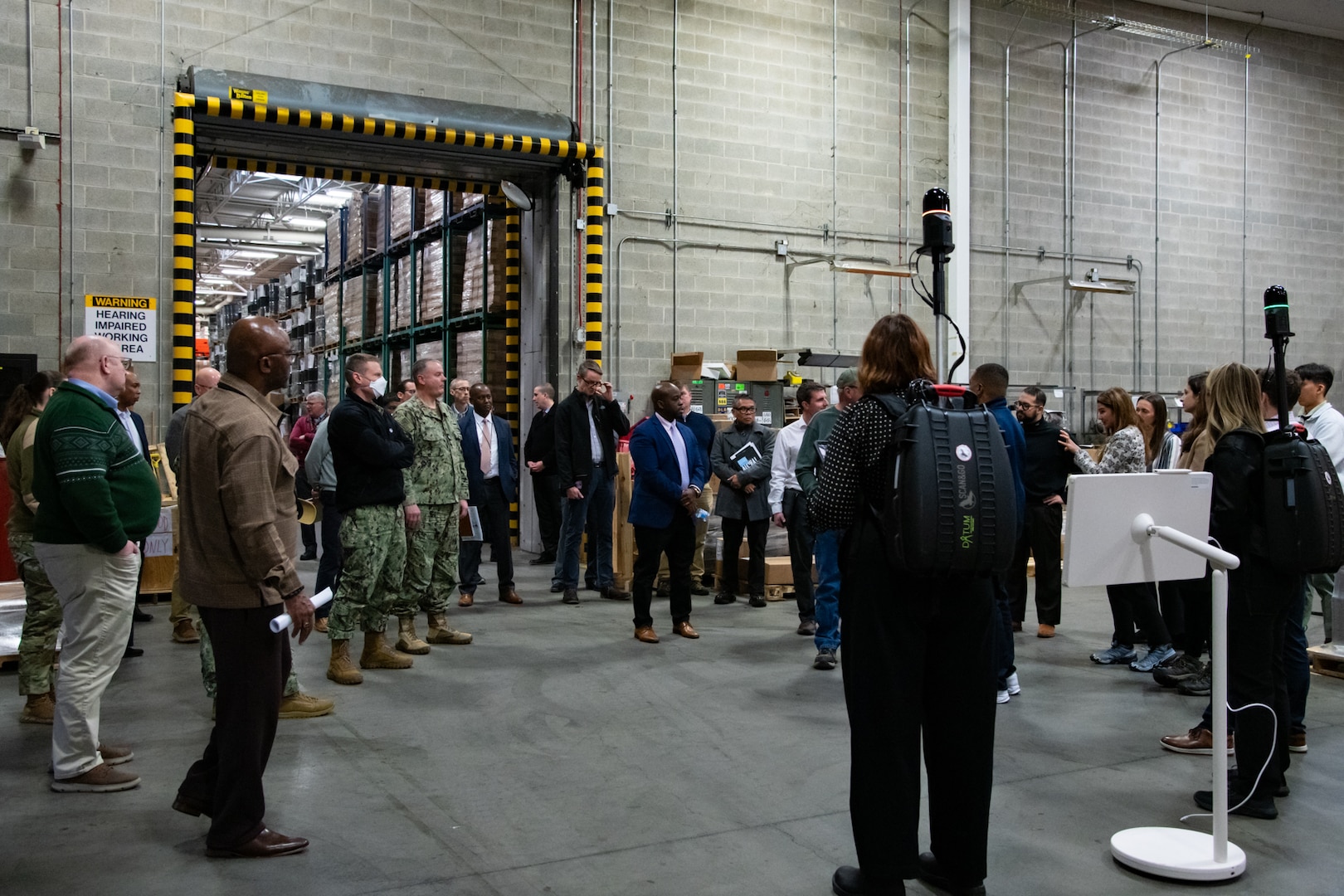 A group of people stand inside a warehouse talking to each other and looking at two people wearing lidar backpacks.