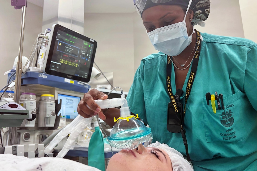 A woman wearing surgical scrubs and head cover places an oxygen mask on a woman lying down wearing a hospital gown and cap. Medical equipment and supplies are in the background.