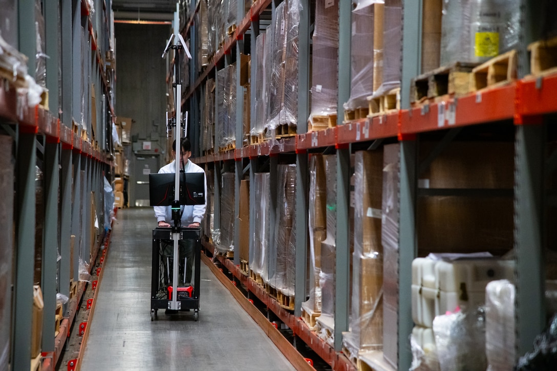 A man walks in between rows of shelving inside of a warehouse pushing a cart with computer equipment on it.