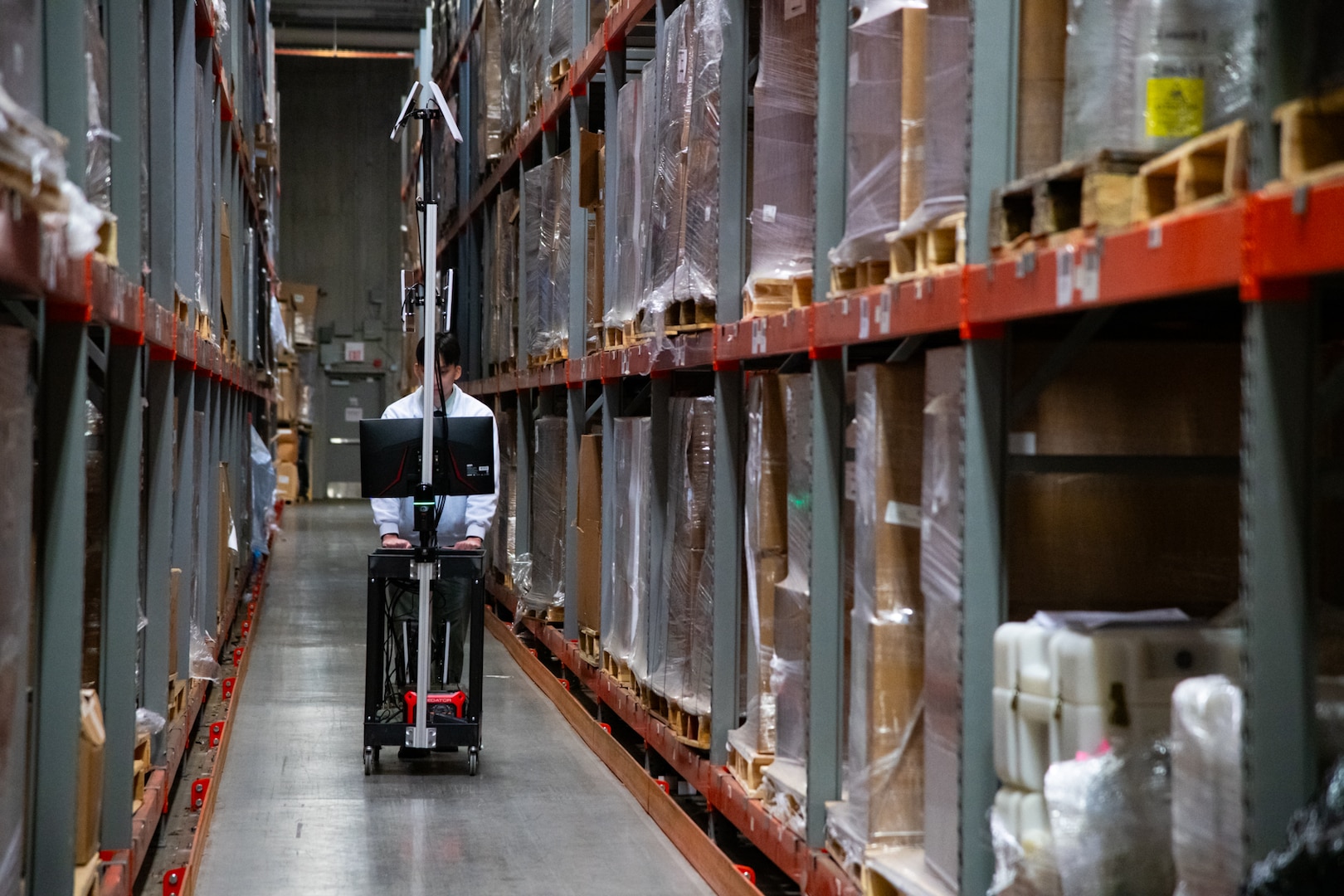 A man walks in between rows of shelving inside of a warehouse pushing a cart with computer equipment on it.
