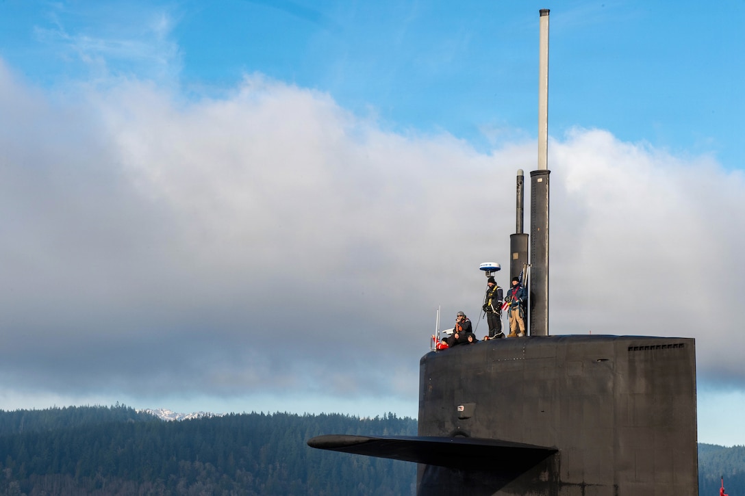 Three sailors are harnessed to the sail of a submarine, as two others visible from the head up also look out, against a wooded area in the background under a cloudy, blue sky.