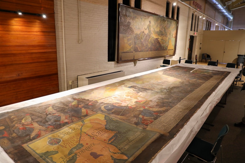 A large painting depicting a battle scene is laid out on tables. A similar painting hangs on a white brick wall in the background.