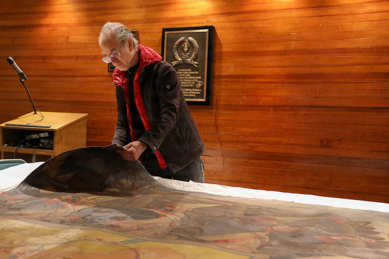 A man wearing glasses and casual attire lifts the corner of an old, large painting to examine it.