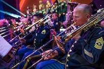 Army band musicians wearing ceremonial uniforms are performing on trombones and various instruments during a concert.