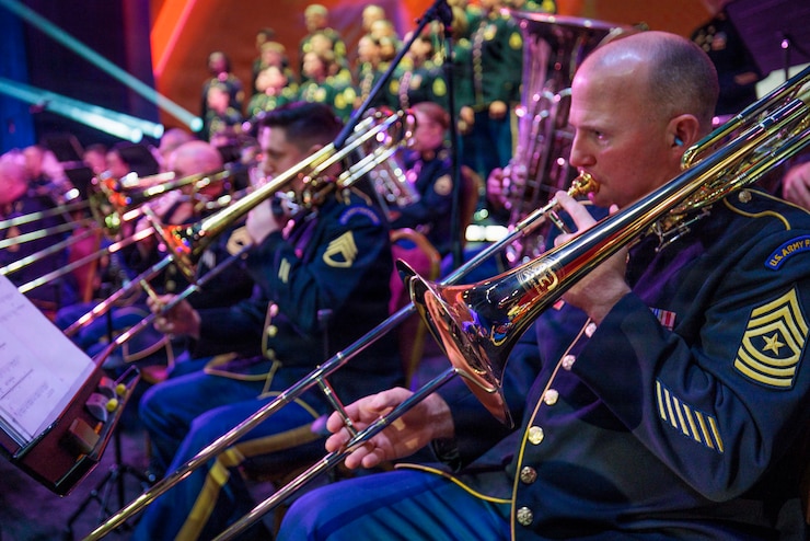 Army band musicians wearing ceremonial uniforms are performing on trombones and various instruments during a concert.