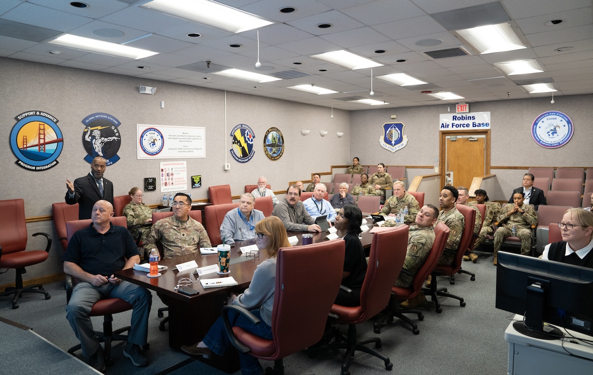 Group of business professionals and Air Force officials sit around table for briefing. Chairs are red.