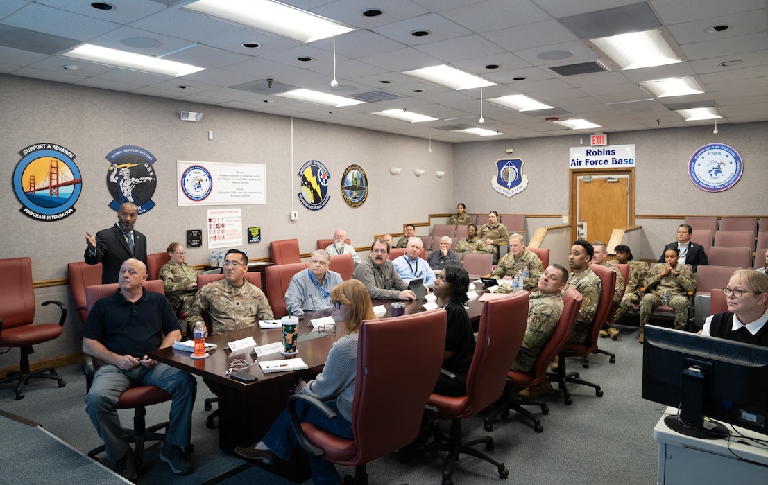 Group of business professionals and Air Force officials sit around table for briefing. Chairs are red.