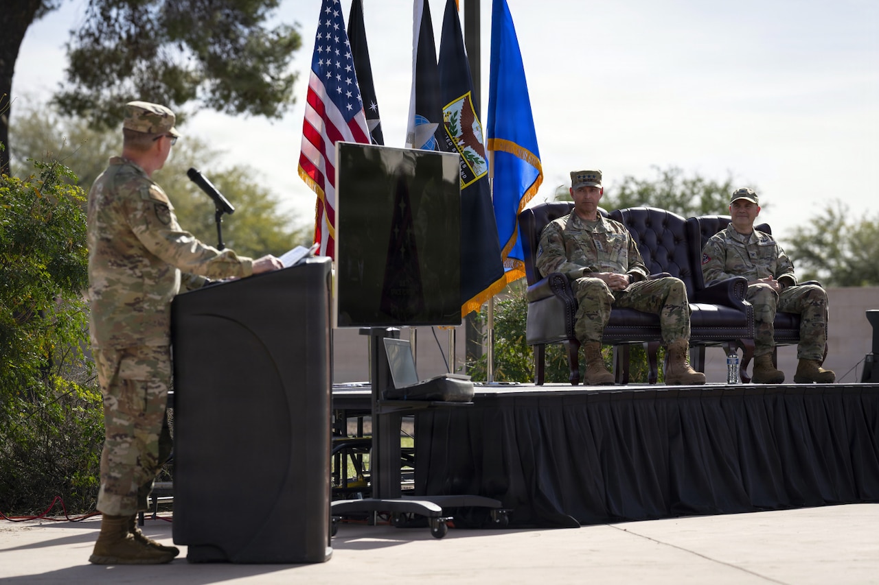 Two men in camouflage military uniforms sit in chairs on a stage outside while looking at another man in similar attire as he speaks into a microphone. There are several flags displayed on the stage next to the men.