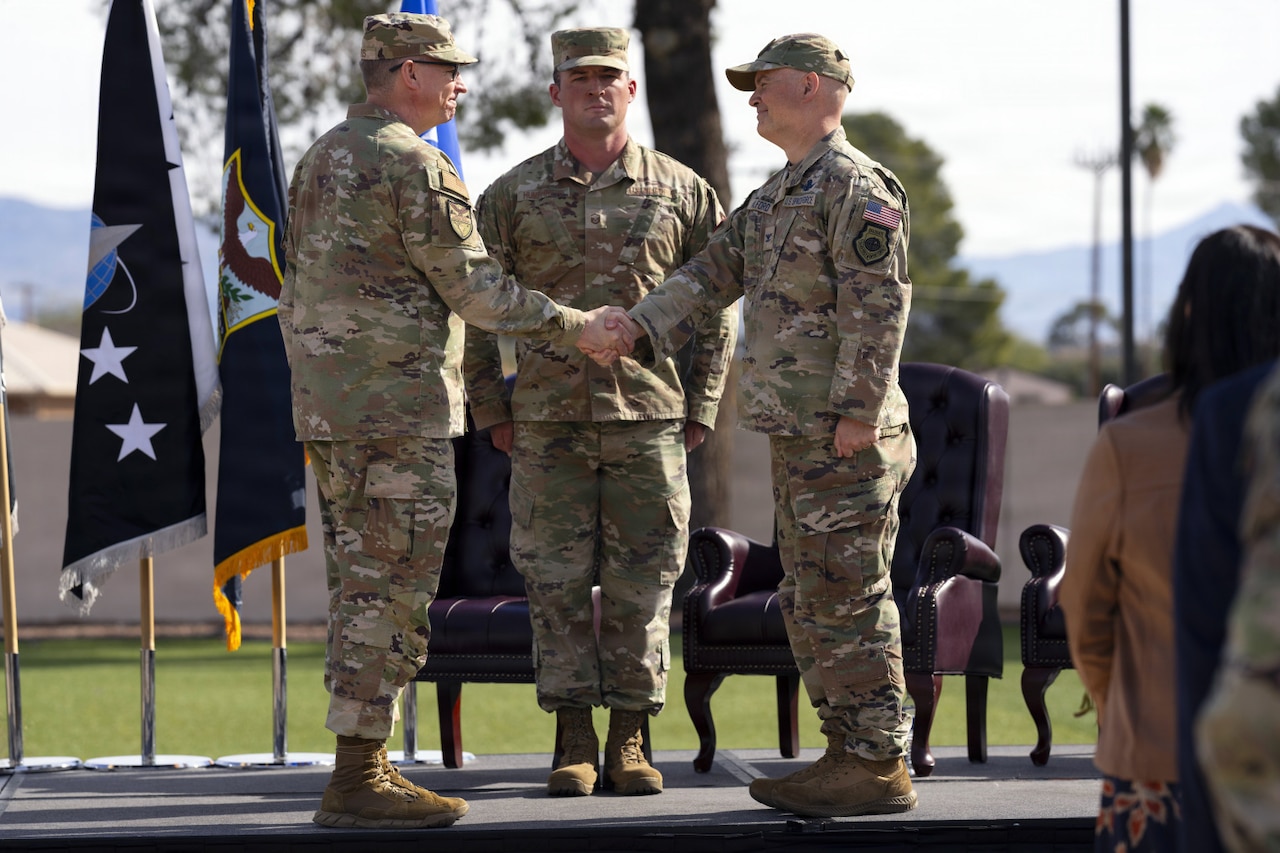 Two men in camouflage military uniforms shake hands while standing on a stage. There is another man in similar attire standing at attention behind them. There are flags displayed and three chairs on stage behind the men.