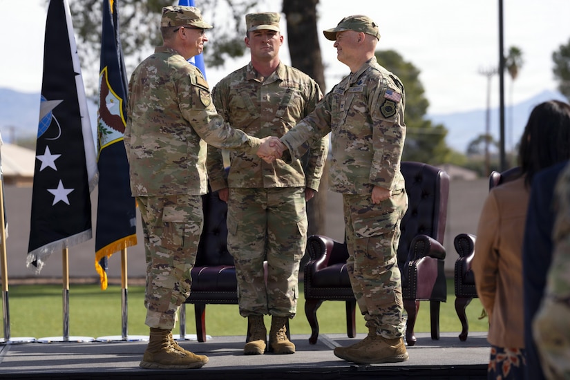 Two men in camouflage military uniforms shake hands while standing on a stage. There is another man in similar attire standing at attention behind them. There are flags displayed and three chairs on stage behind the men.