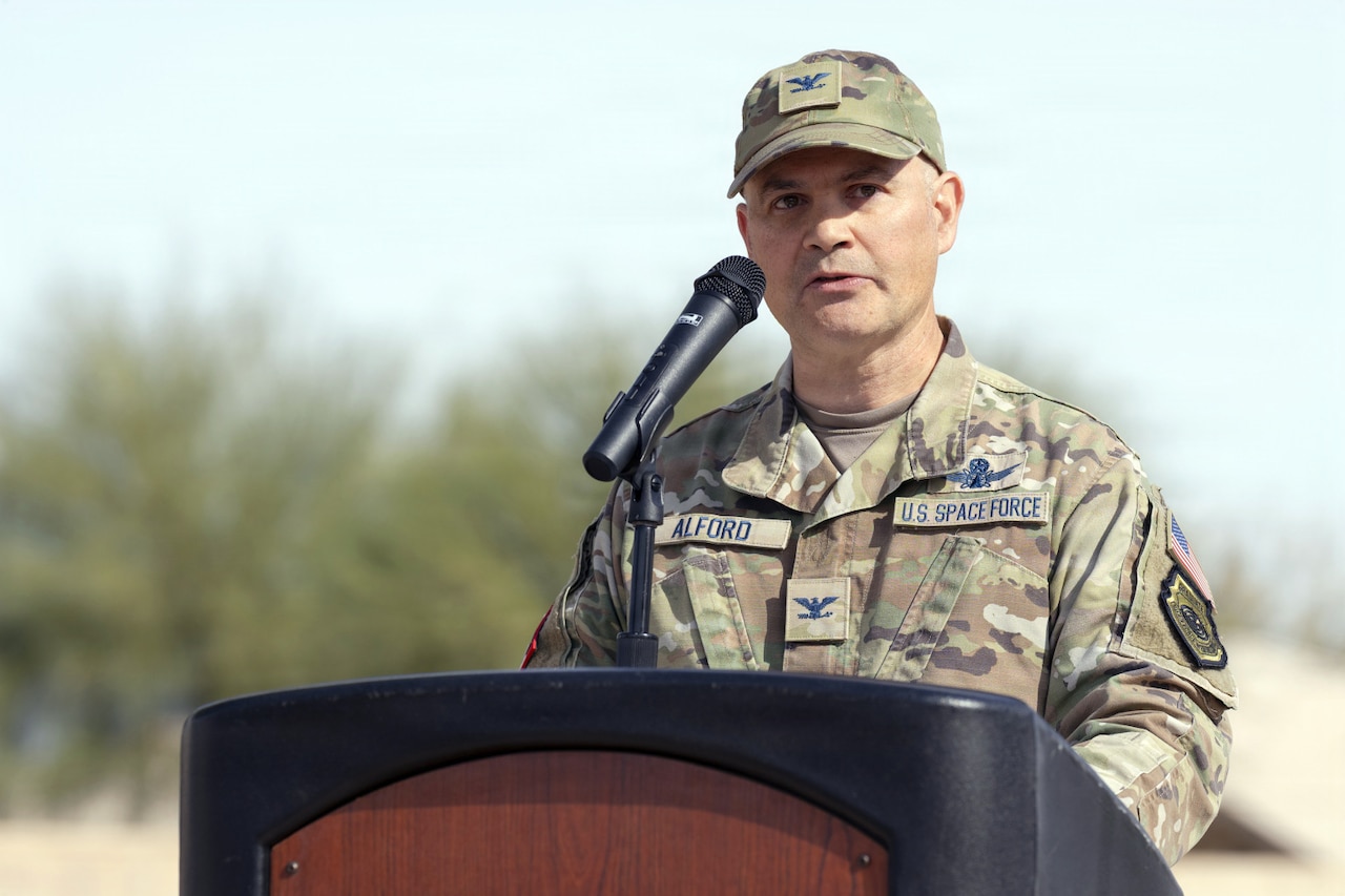 A man in a camouflage military uniform speaks into a microphone while standing behind a lectern.