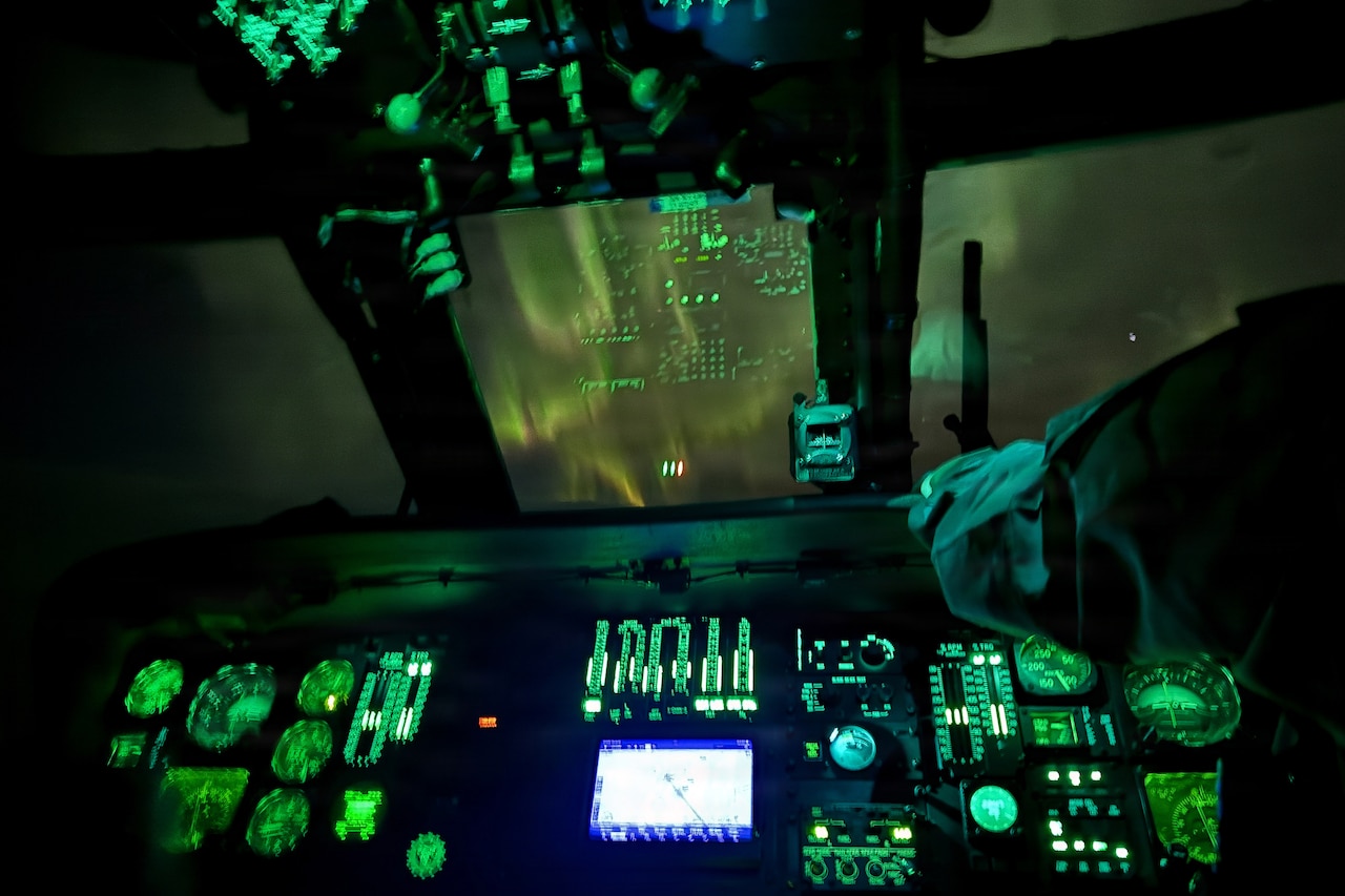 A helicopter pilot wearing a military uniform reaches his arm up toward the cockpit. In the night sky, a ribbon of soft colorful lights can be seen.