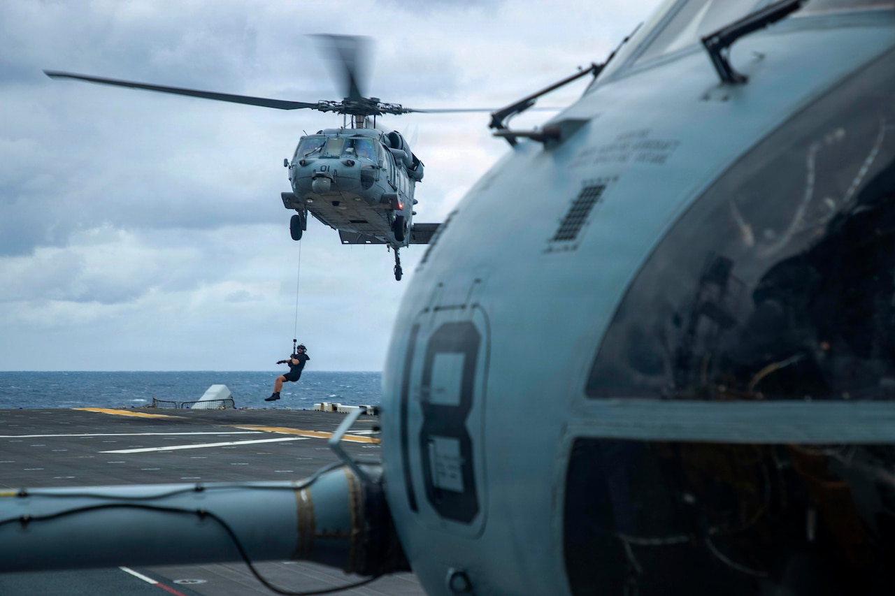 A man rappels from a helicopter as it lands on the flight deck of a ship at sea; a part of another helicopter is seen on the right in the foreground.