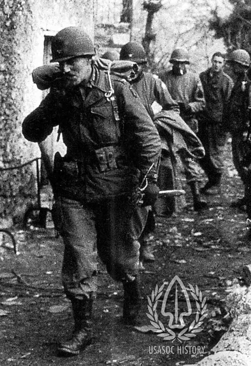 A group of men in military combat gear walk single file along a dirt path.