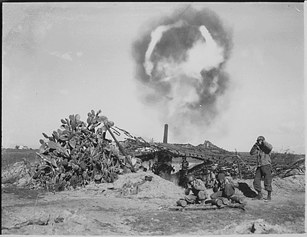 A long gun aimed skyward lets off a plume of flames. Two men in military uniforms kneel as a third man in similar attire stands while covering their ears. Foliage can be seen around them in a barren landscape.