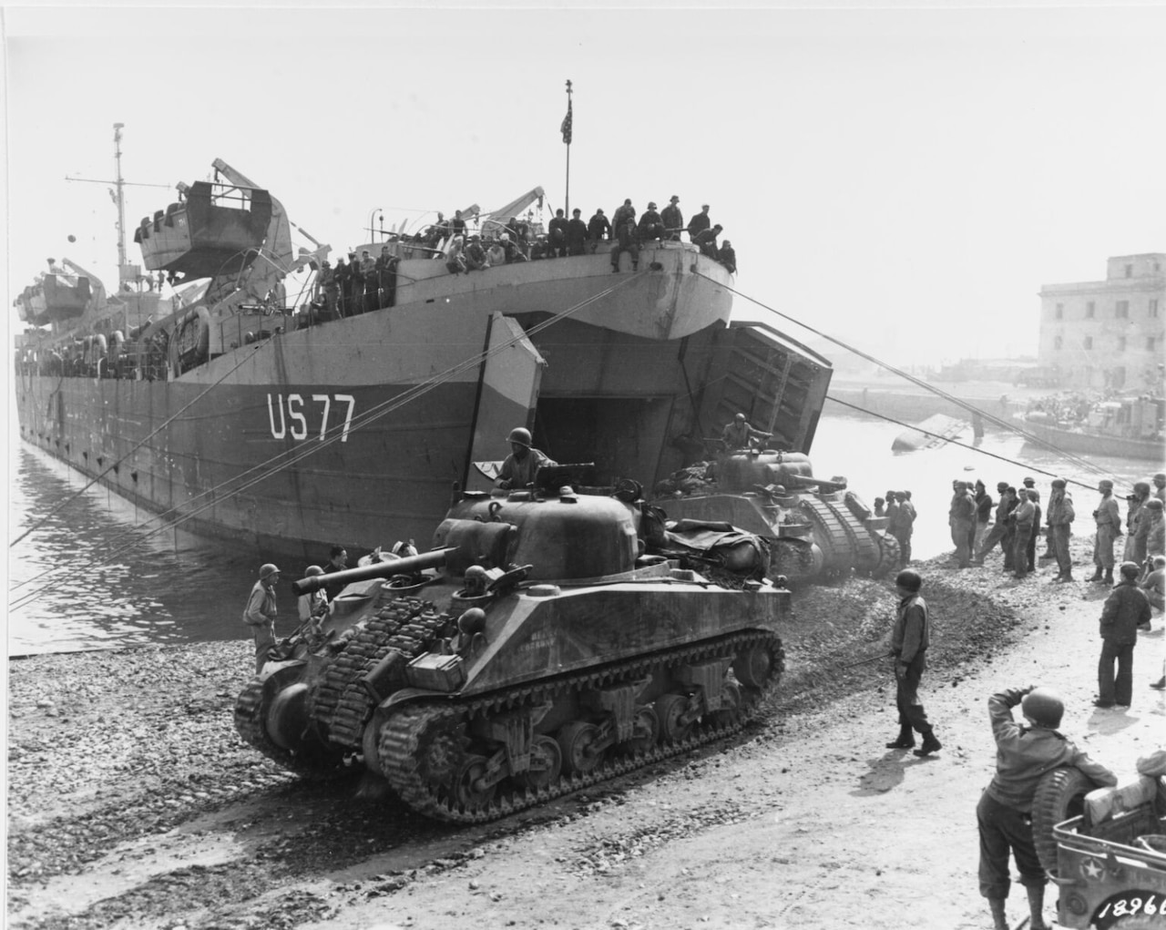 A large ship with ropes coming from its bow and an open front sits on a beach. In front of it sits a large tank as another one rolls off the ship. Dozens of men in military uniforms stand on the ship while several others in similar attire stand on the beach.