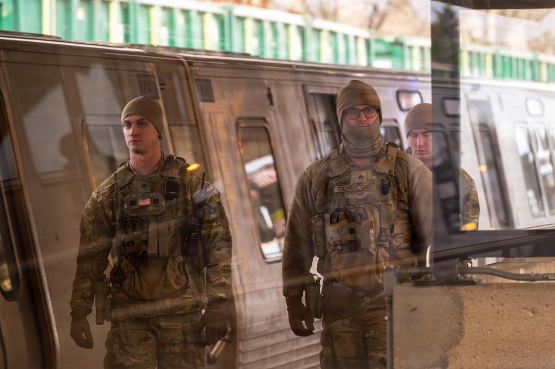 Three men wearing camouflage military uniforms and head covers are seen through a glass partition as they walk past a metro train.