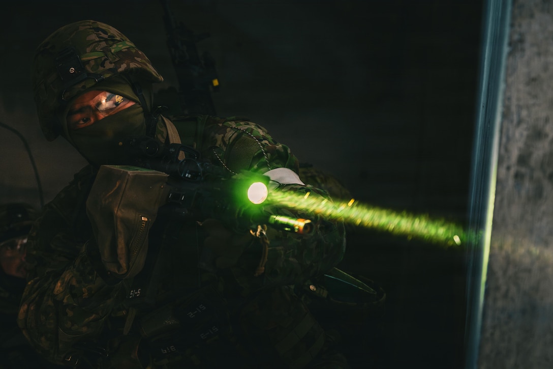 A person wearing a camouflage military uniform and head cover holds a weapon in the dark; a bright neon glow is coming from the open end of the nozzle.