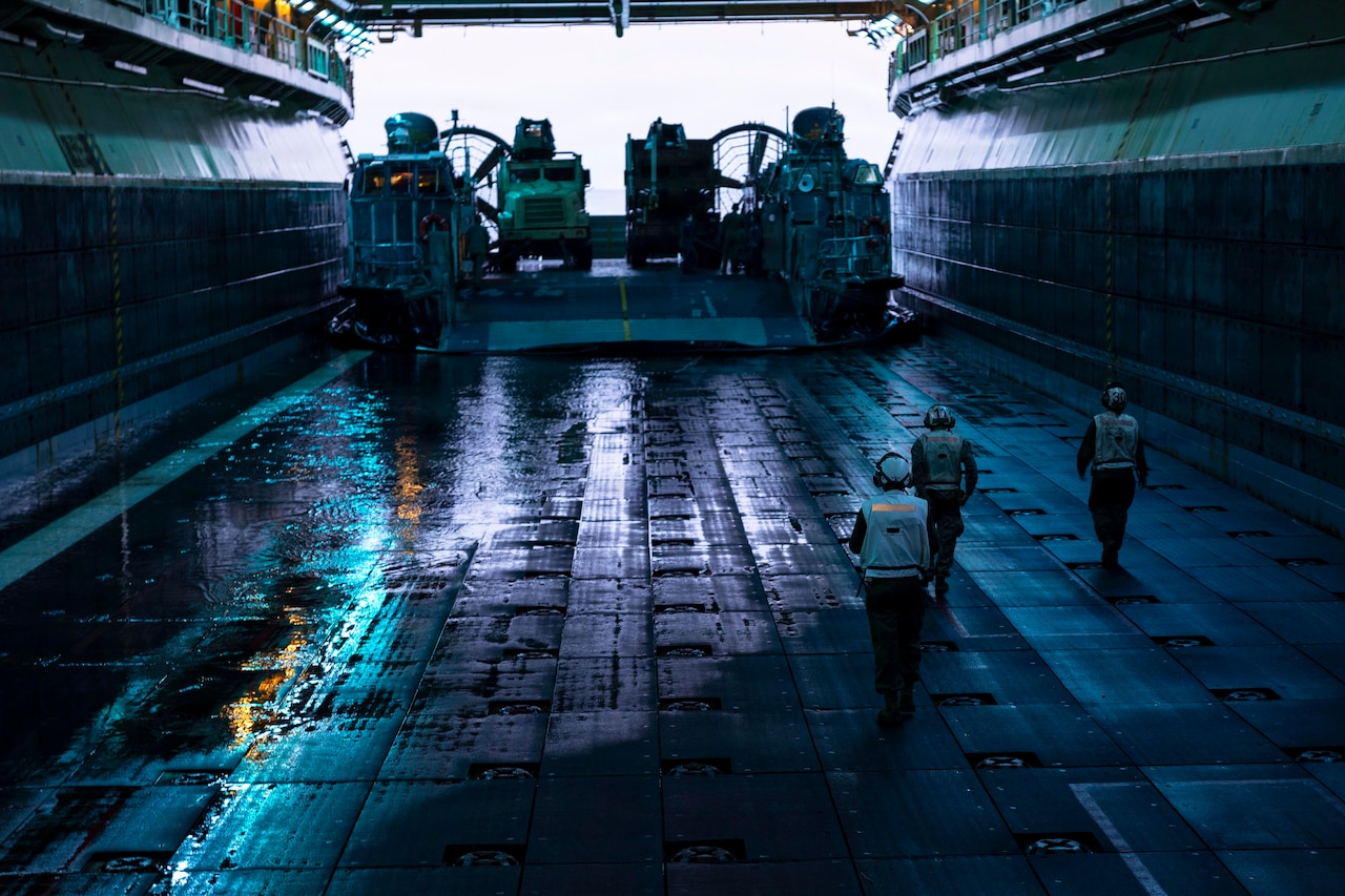 Three people in a dark well deck wearing flight gear and headsets walk toward a landing craft transporting trucks.