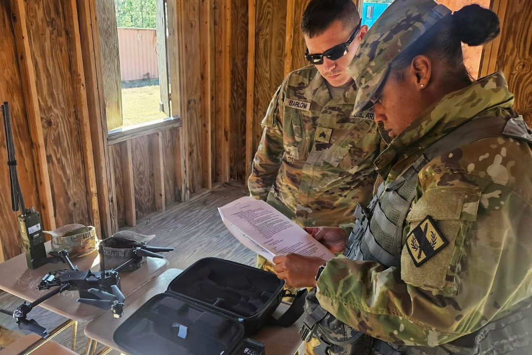 Two guardsmen inside a wood structure stand and look at paperwork, with a table nearby that has a small drone and other electronic equipment on it.