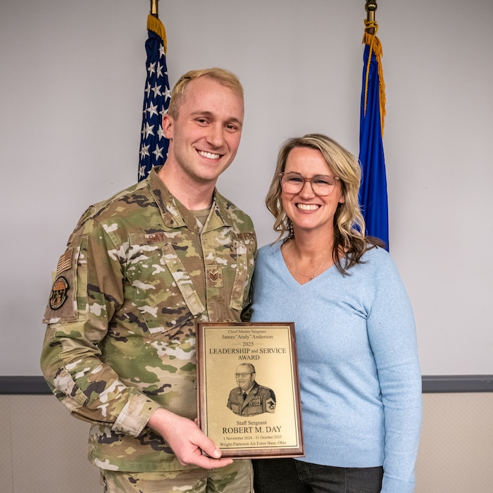 A blonde man, wearing military camouflage uniform, poses with a blonde woman, wearing glasses and light blue shirt, while holding an award plaque