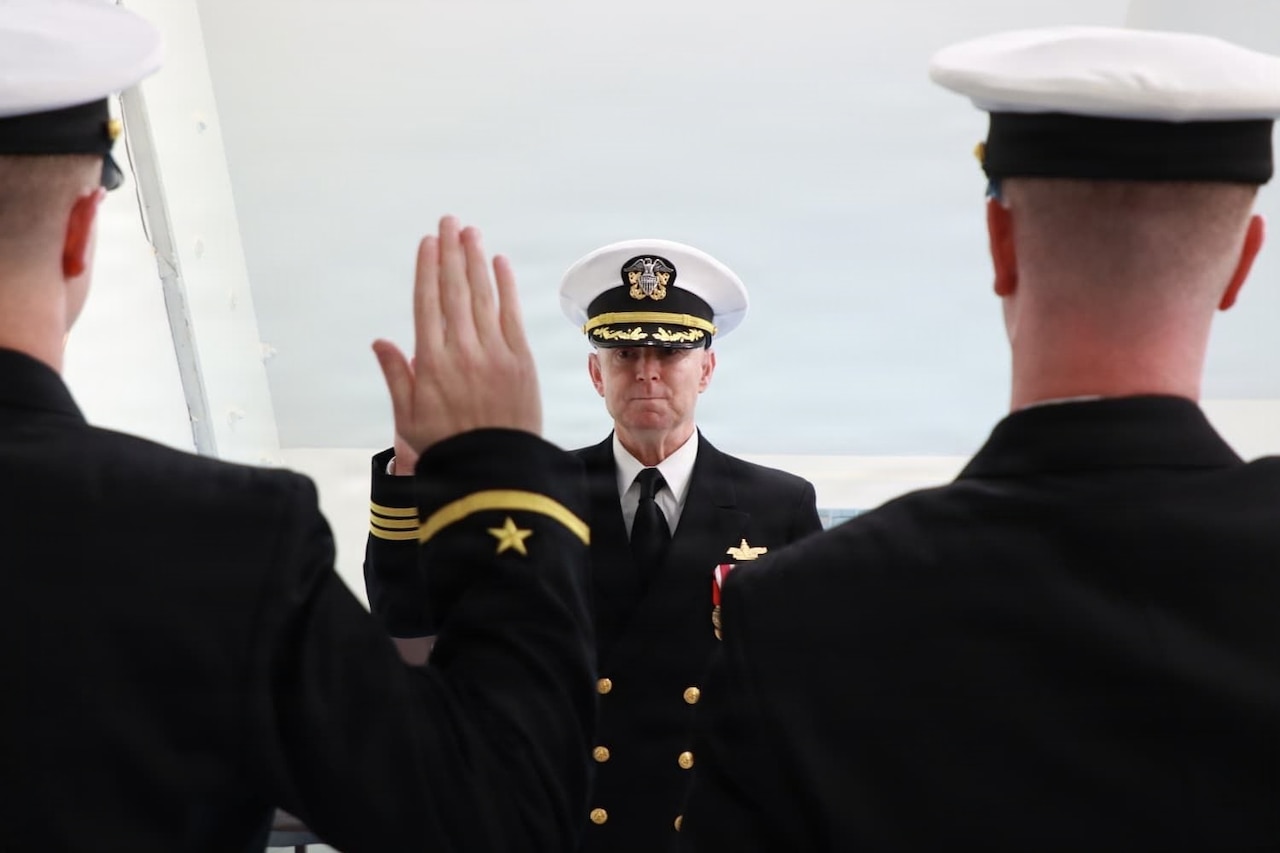 A U.S. Navy officer administers the oath of office to two other officers; all three men are in dress uniforms with their right hands raised.