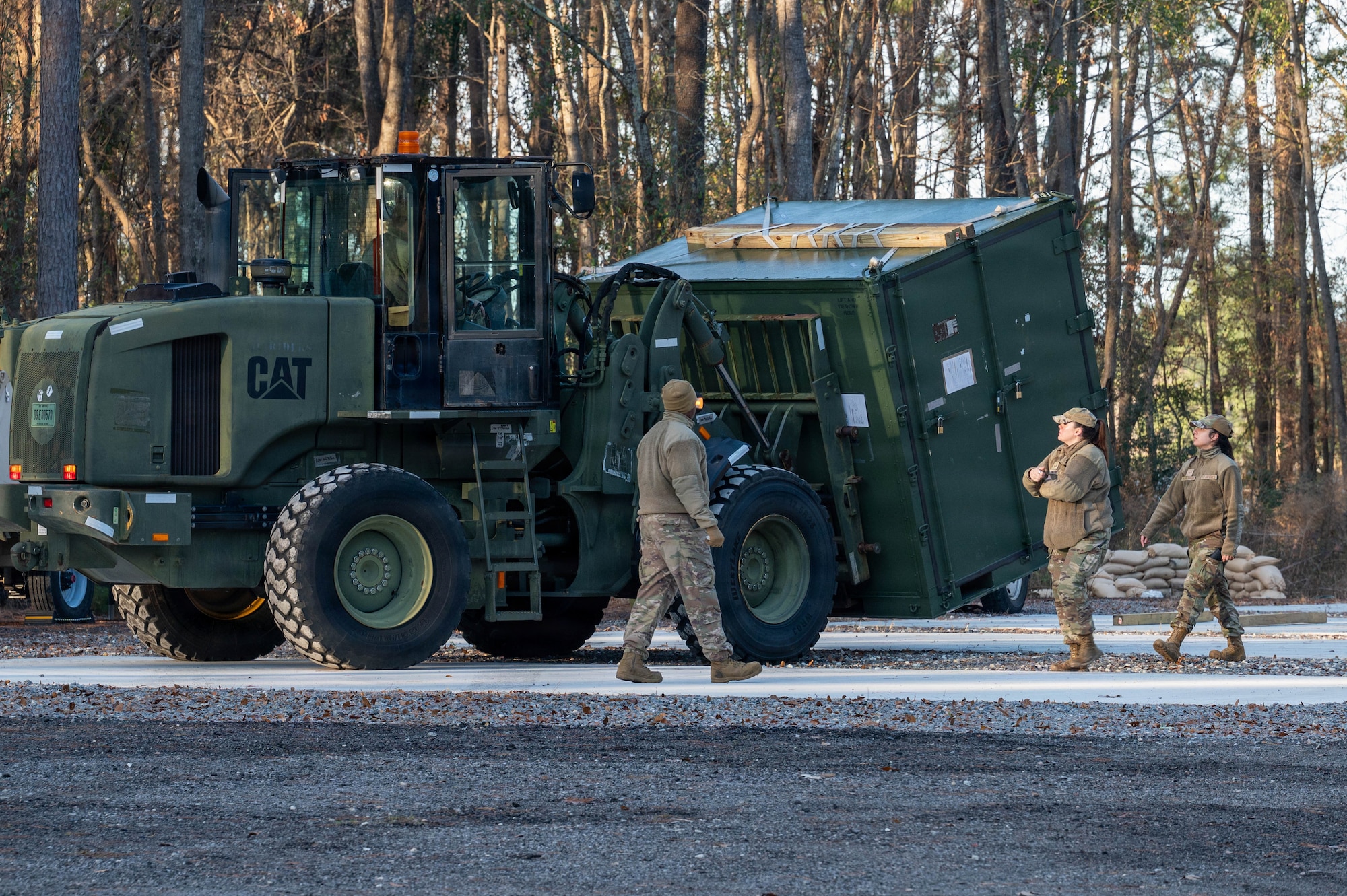 A forklift moves a storage container across a clearing while three Airmen supervise.