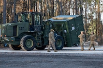A forklift moves a storage container across a clearing while three Airmen supervise.