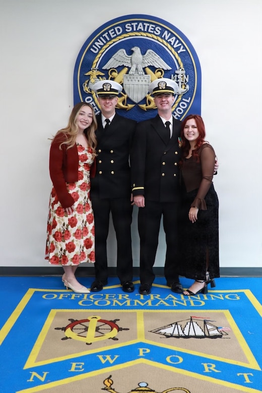 Two men in U.S. Navy uniforms and their wives smile for a group photograph in front of a naval seal.