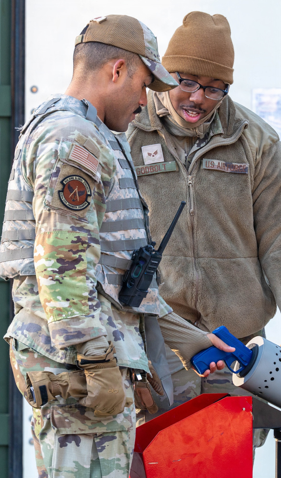 An Airman stands with a blue plastic weapon into a clearing chamber while another supervises.